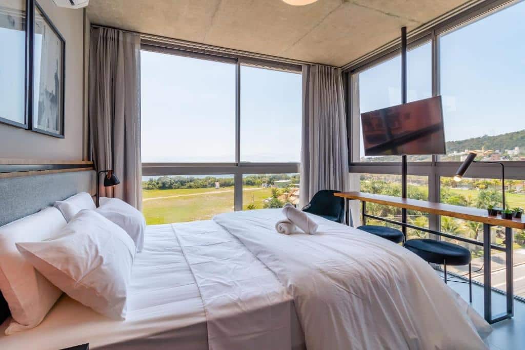 Quarto do airbnb Spot Jurere sofisticação à beira mar. A direita há uma televisão suspensa presa a um poste fino que vai do chão ao teto, abaixo há uma bancada de madeira com duas cadeiras e um abajur, além de uma poltrona ao lado. Ao centro é possível ver a cama de casal, que fica de frente para a televisão. As paredes da direita e do fundo são inteiramente de vidro, contendo cortinas para cobri-las. Imagem utilizada para ilustrar o post airbnb em Jurerê Internacional.
