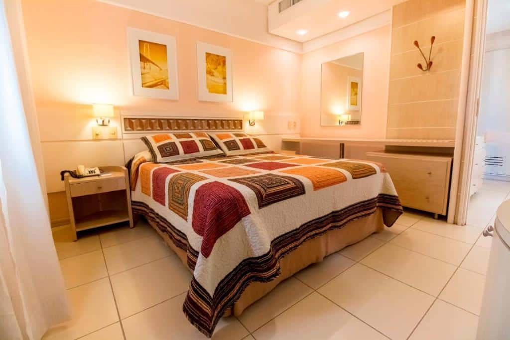 Quarto do airbnb Suítes e Studios no Hotel Jurerê Beach Village. No centro do quarto há uma cama de casal, em cada lado da cama há uma mesa de centro com um abajur e no lado direito da cama há uma prateleira extensa presa a parede. Imagem utilizada para ilustrar o post airbnb em Jurerê Internacional.