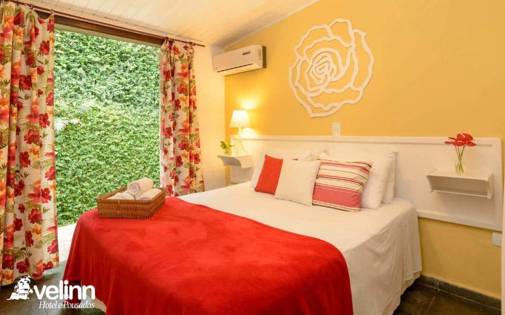 Quarto da VELINN Caravela Pousada. Há uma cama de casal com prateleiras enfeitadas de ambos os lados. Ao fundo pode-se ver uma porta com cortinas, que vai para a área externa. Imagem utilizada para ilustrar o post pousadas pet friendly em Ilhabela.