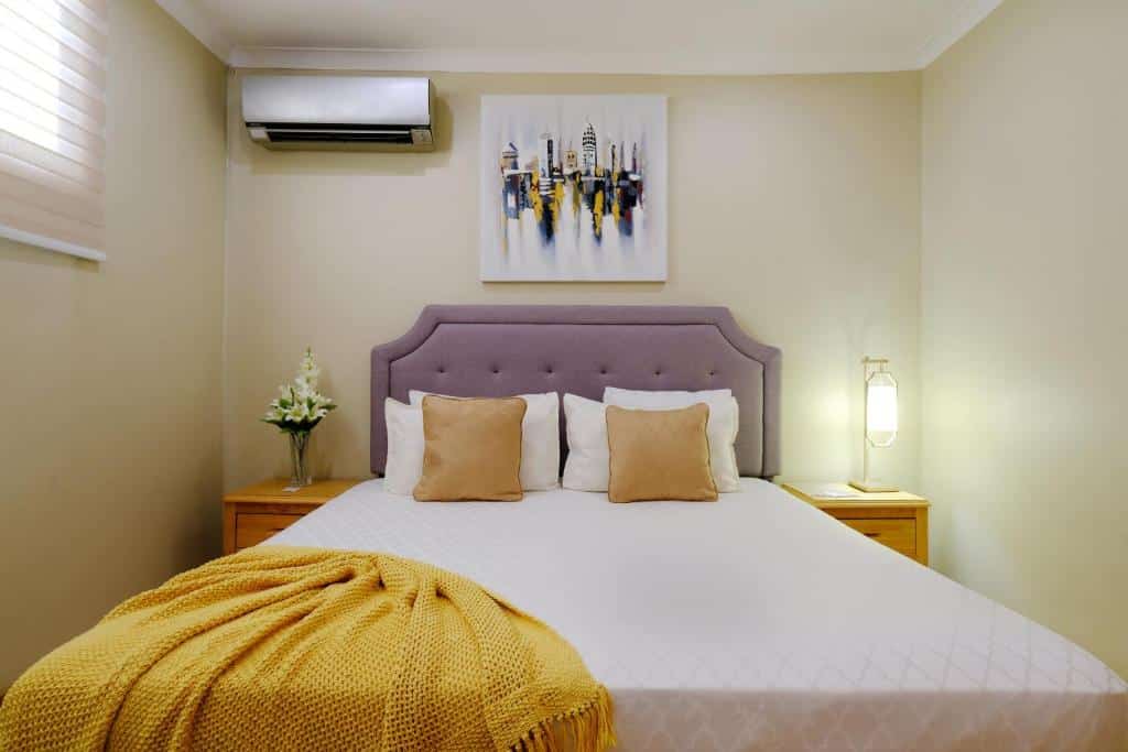 Quarto do Victoria City Hotel com cama de casal no centro da imagem em cada lado da cama uma cômoda do lado direito um vaso de flor em cima da cômoda e do lado esquerdo uma luminária. Representa Aruba.