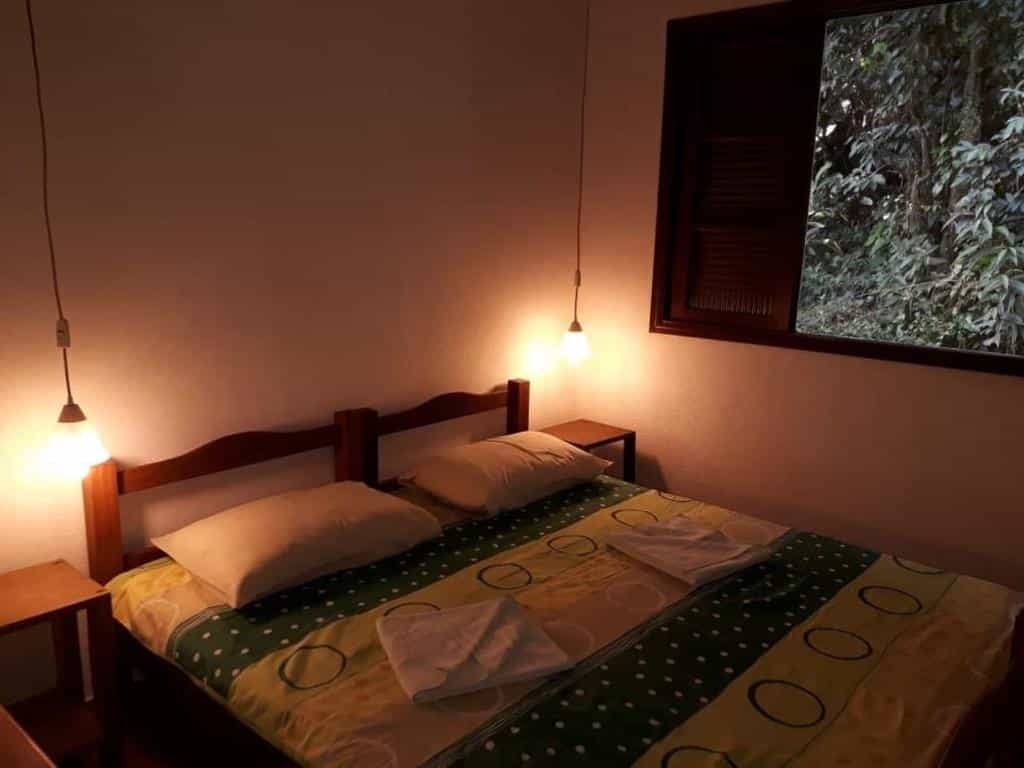 Quarto da Vila Betânia que mostra uma cama de casal com dois travesseiros, duas mesas de cabeceira de cada lado da cama e duas luminárias penduradas. Do lado direito da cama tem uma janela aberta com vista para a paisagem.