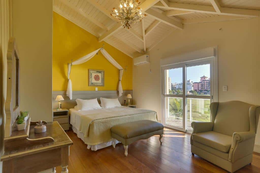 Quarto do airbnb Villas Jurerê Residences. A direita há uma grande poltrona e a esquerda uma penteadeira com espelho. Ao fundo há uma cama de casal centralizada com cômodas e abajures de ambos os lados. Do lado direito da cama há uma porta de vidro que vai para a varanda do quarto. Imagem utilizada para ilustrar o post airbnb em Jurerê Internacional.