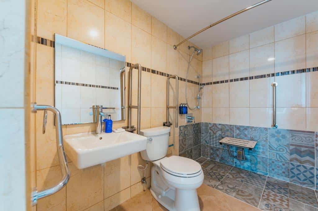 Banheiro com acessibilidade do Radisson Blu Aruba. Do lado direito uma pia e um vaso sanitário com diversas barras, no fundo o box com acento e a barras.