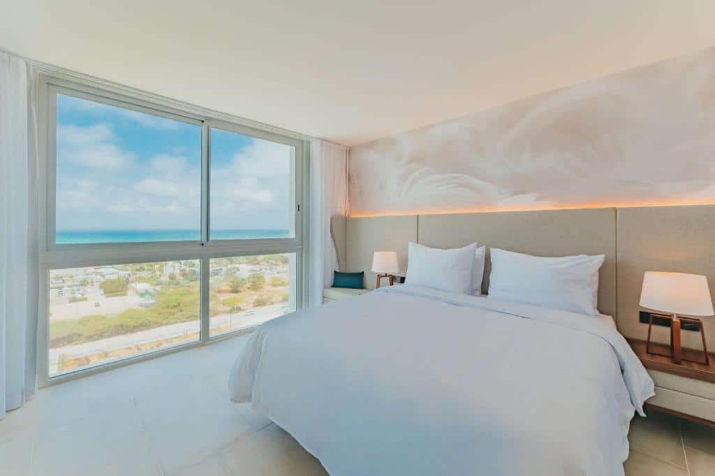 Quarto do Radisson Blu Aruba. Uma cama de casal do lado direito, de cada lado um abajur. Do lado esquerdo a parede e janela de vidro com vista para o mar.