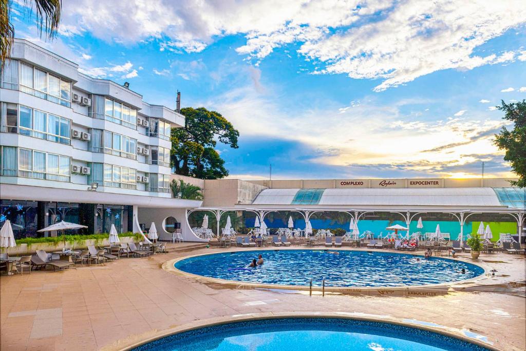 Parte do Rafain Palace Hotel & Convention Center que mostra a área da piscina com duas psicinas redondas, algumas espreguiçadeiras e guarda-sóis em volta. Ao fundo tem uma área coberta e do lado esquerdo tem a construção do hotel. Imagem ilustrando post Hotéis em Foz do Iguaçu.