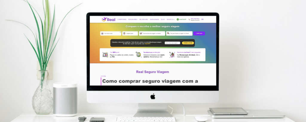 Um computador sob uma mesa branca com alguns itens de escritório e um vaso de planta, na tela está aberto o site da Real Seguro Viagem