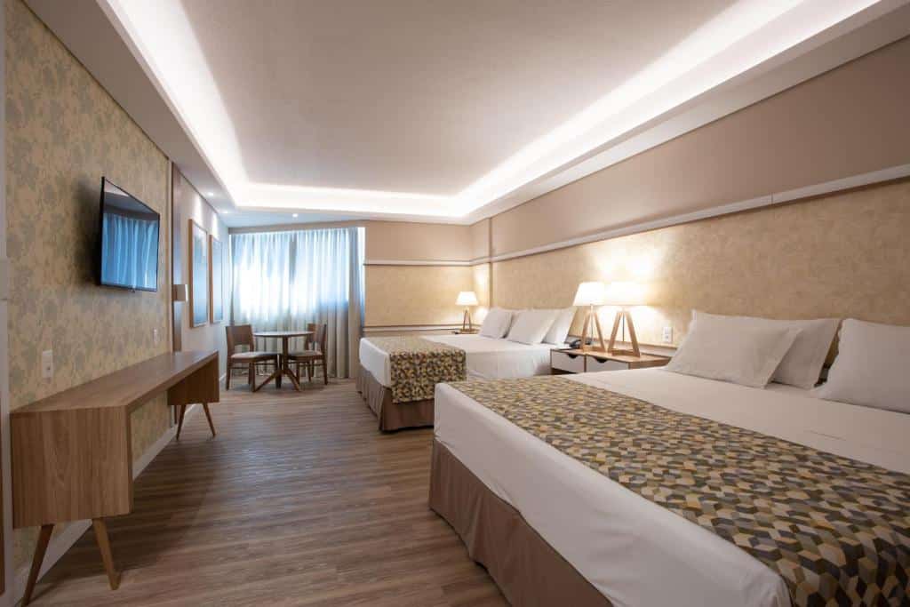 Foto do quarto do Recanto Cataratas – Thermas, Resort e Convention, ilustrando o post sobre Onde ficar em Foz do Iguaçu. Há duas camas box de casal na direita, e entre elas há uma mesa de cabeceira com abajur em cima. Na frente delas há uma mesa e uma TV. No fundo do quarto há uma janela e uma mesa redonda com duas cadeiras.