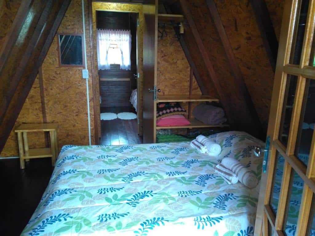 Imagem do quarto em Recanto Mauá - Chalé Azaleia, que está ilustrando o post sobre airbnb em Visconde de Mauá. Há uma cama de casal, e do seu lado direito há uma porta com varanda, e do outro lado há a porta do quarto e umas prateleiras para guardar roupas e cobertores.