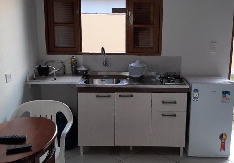 Cozinha do Recanto Primavera. Uma pia com armário e fogão está a lado de um frigobar. Em frente é possível ver uma mesa e uma cadeira.