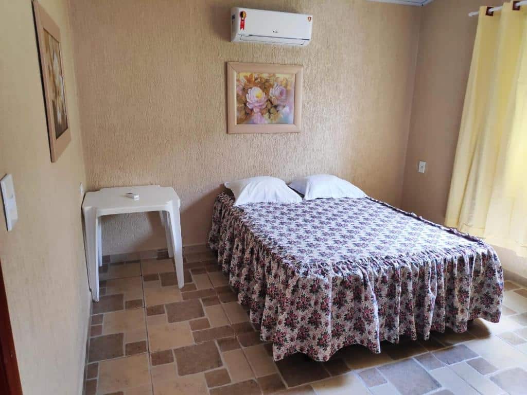 Quarto de uma das casas do Recanto Primavera Ubatuba. Uma cama de casal está no meio, ao lado de uma mesa de plástico e uma janela. Na parede, acima dela, está um ar-condicionado. Esse é um dos airbnb na Praia do Sapê.