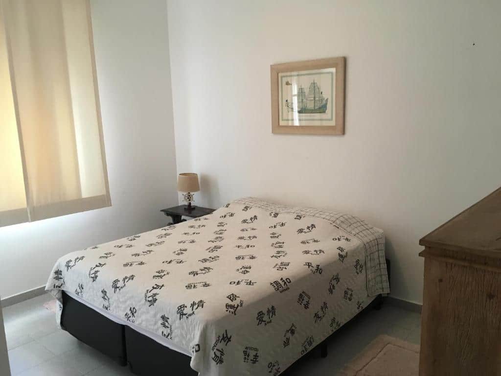 Imagem do quarto do Relax GJA Frente ao Mar, ilustrando o post sobre airbnb em Guarujá. Há uma cama de casal box no centro, e ao lado dela há uma mesa de cabeceira com abajur. Na esquerda há uma janela. No canto inferior direito vemos o relance de uma cômoda.