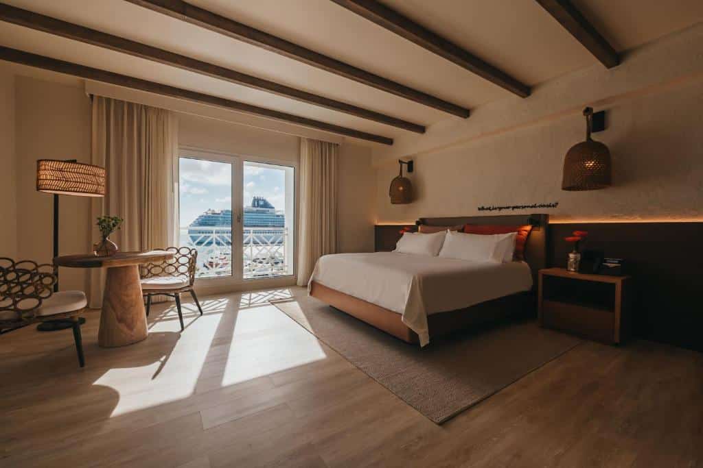Quarto do Renaissance Wind Creek Aruba Resort. Uma cama de casal está no canto, perto de uma porta de vidro levanda até a varanda e com visão para o mar. Em frente a cama es[ta uma mesa com duas cadeiras. Esta é uma imagem para o post de hotéis de luxo em Aruba.