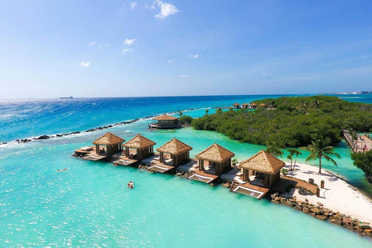 Cabanas do Renaissance Wind Creek Aruba Resort na ilha privativa. Ela estão em uma curta faixa de areia no meio do mar. Ao fundo é possível ver diversas árvores.
