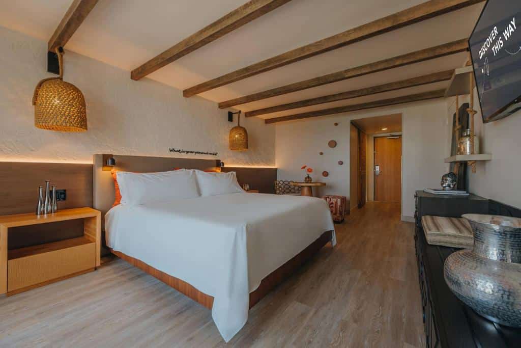 Quarto do Renaissance Wind Creek Resort. Uma cama de casal do lado esquerdo com uma cômoda e um lustre de cada lado, no fundo uma mesinha. Do lado direito uma raque com decorações e uma tv. Foto para ilustrar post de onde ficar em Aruba.