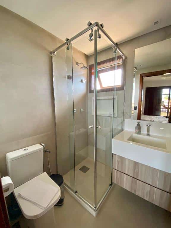 Banheiro moderno, limpo e com cores claras em tons de branco e bege no Residencial Bali Bahia Apto Alto Padrão BATUR 106. Há uma privada, chuveiro com box de vidro e pia com espelho.