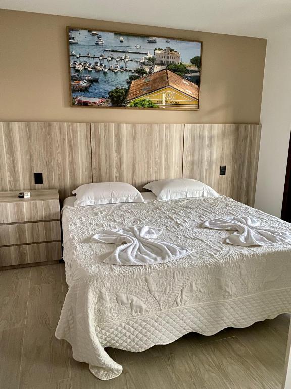 Foto do quarto em Residencial Bali Bahia Apto Alto Padrão BATUR 106, que ilustra o post de airbnb na Praia do Forte. Tem uma cama box de casal no centro, com cabeceira atrás de madeira que também conta com uma mesa de cabeceira ao lado esquerdo da cama. Na parede há um quadro da Praia do Forte.