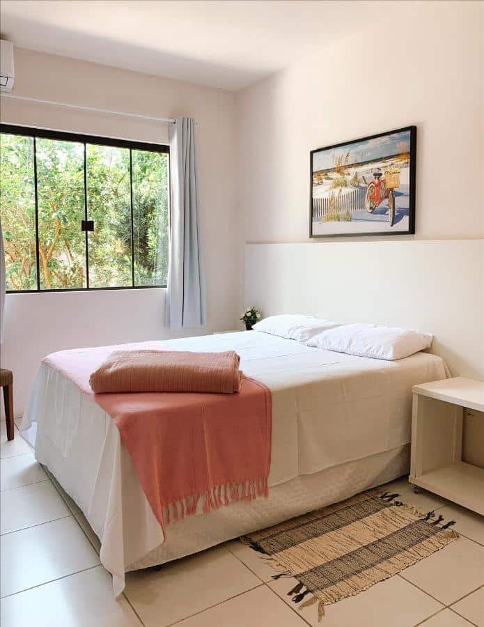 Quarto do Residencial COLIBRI, airbnb em Garopaba. Uma cama de casal está encostada numa cabeceira branca na parede direita com mesinhas dos dois lados. Há um quadrinho na parede acima da cama, e ao lado esquerdo fica uma janela de vidro com cortinas e vista para árvores.
