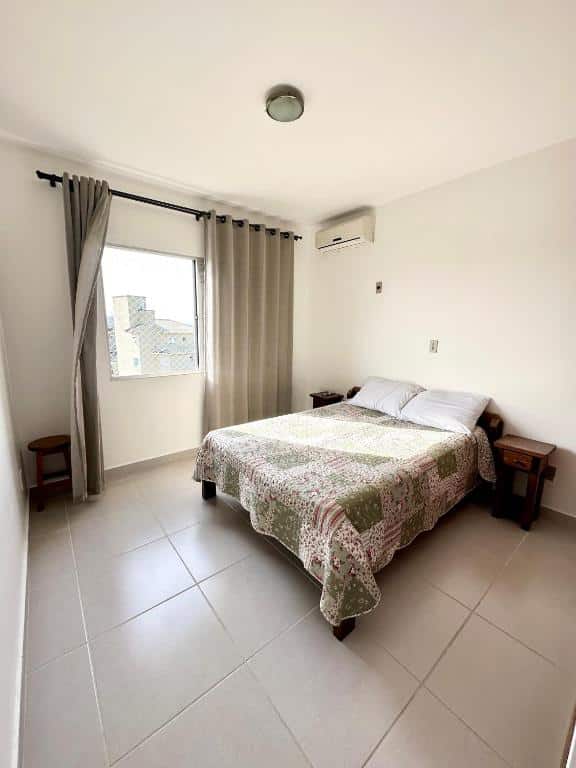 Quarto do airbnb Residencial Mansur. No centro do quarto há uma cama de casal, com cômodas dos dois lados. Na esquerda têm uma janela com tela de proteção e uma cortina. Imagem utilizada para ilustrar o post airbnb em Jurerê Internacional.