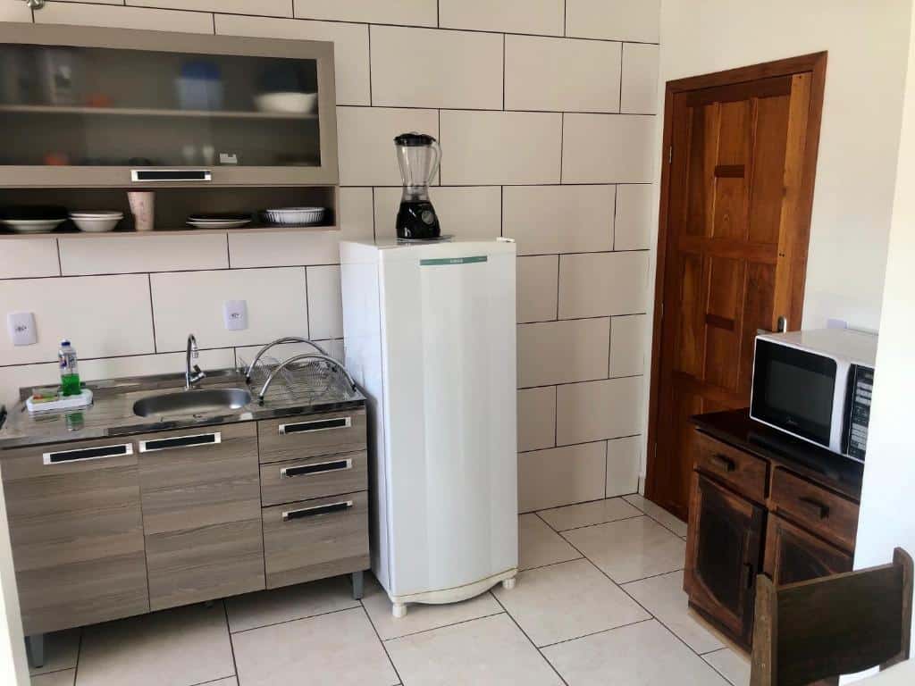 Cozinha do Residencial Sandini Garopaba. Ao lado esquerdo está a bancada da pia e o armário suspenso com utensílios. No centro fica a geladeira com um liquidificador acima. Na lateral direita há uma porta, e ao lado fica um móvel de madeira com um micro-ondas em cima.