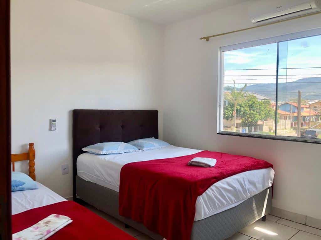Quarto do Residencial Sandini Garopaba, airbnb em Garopaba. Uma cama de casal está encostada na parede ao lado direito, com uma janela na lateral. Ao seu lado esquerdo fica uma cama de solteiro.