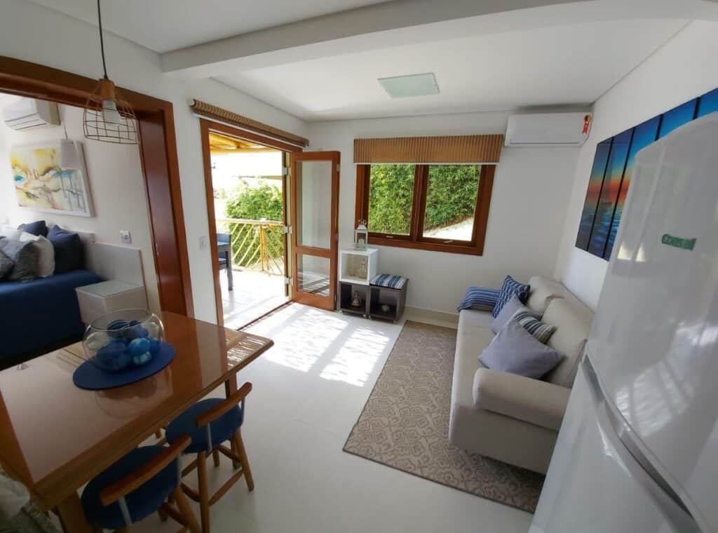 Visão geral do Residencial Yacamim B39. A foto foi tirada entre a geladeira e a mesa de jantar, e ao fundo está a sala de estar com sofá com almofadas e aparelho de ar-condicionado. Uma porta ao lado da mesa de jantar leva ao quarto.