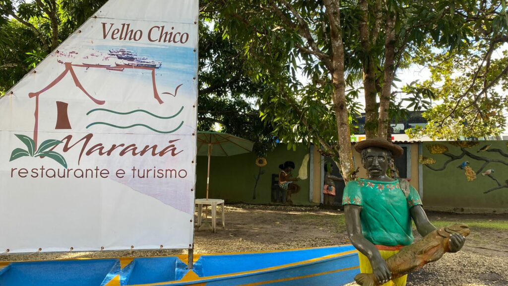 Escultura de um barco de vela escrito na vela "Velho Chico, Maraná Restaurante e Turismo", com um boneco negro ao lado. Há árvores atrás fazendo sombra no ambiente. É nesse restaurante que sai o passeio à Foz do Rio São Francisco