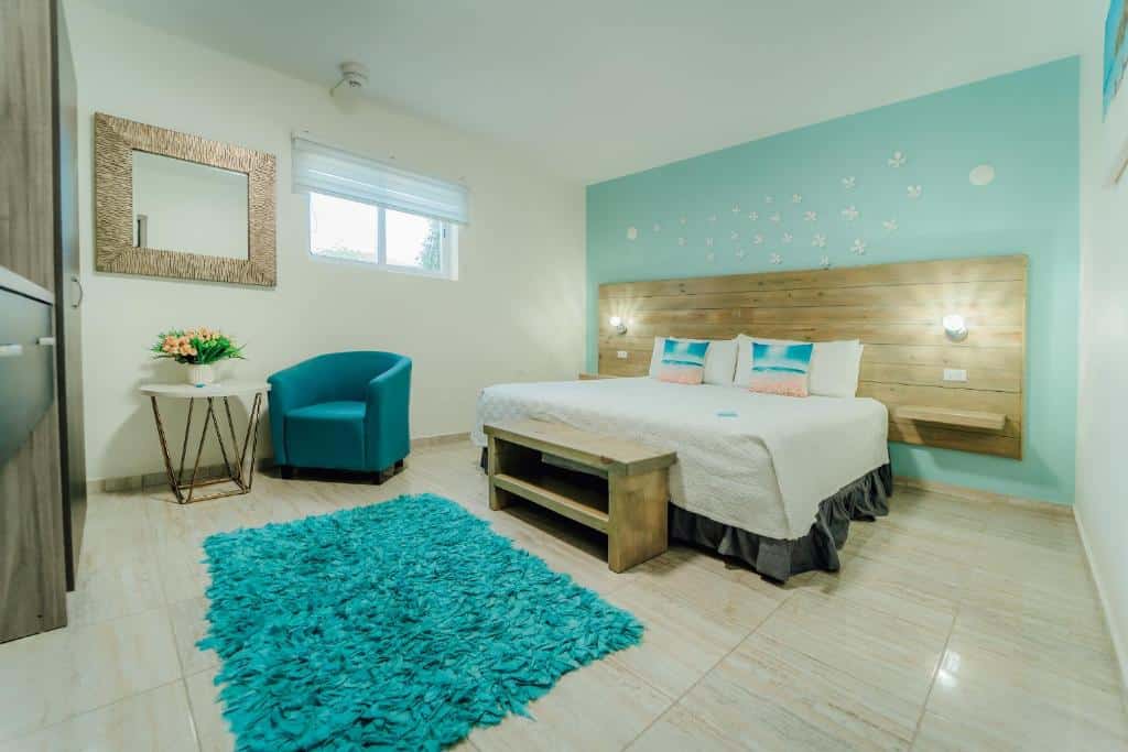 Quarto do RH Boutique Hotel Aruba. Uma cama de casal está ao fundo, com a cabeceira encostada no meio da parede. Na sua ponta esquerd está uma poltrona e uma mesinha, além de um espelho pendurado na parede. Ao pé da cama há um banquinho e logo em seguida um tapete.