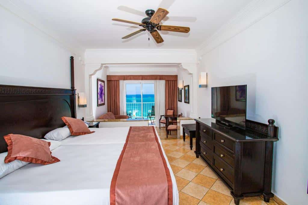 Quarto do Riu Palace Aruba - All Inclusive. Uma cama de casal do lado esquerdo, no meio um ventilador de teto, do lado direito uma cômoda e uma televisão. No fundo uma sala de estar, a varanda e a vista do mar.