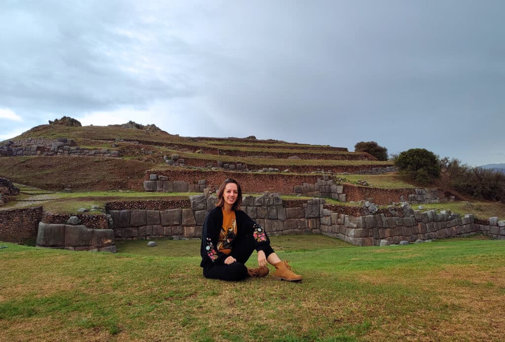 mulher sentada na grama no centro da imagem, atrás dela é possível ver uma construção na terra no sítio arqueológico de sacsayhuaman em Cusco. O dia está nublado e ela usa um casaco de lã preto