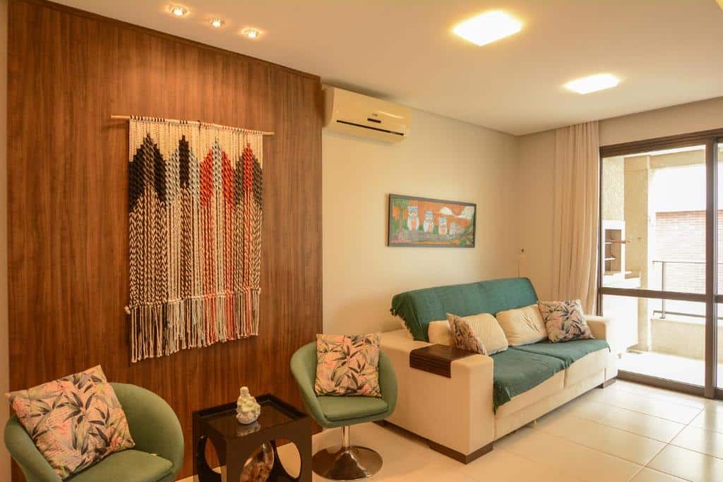 Sala de estar do airbnb Apt Jurerê. Do lado direito há um sofá, que está do lado de uma porta que dá acesso a parte externa da acomodação. No lado esquerdo há duas poltronas e uma mesa decorativa.
