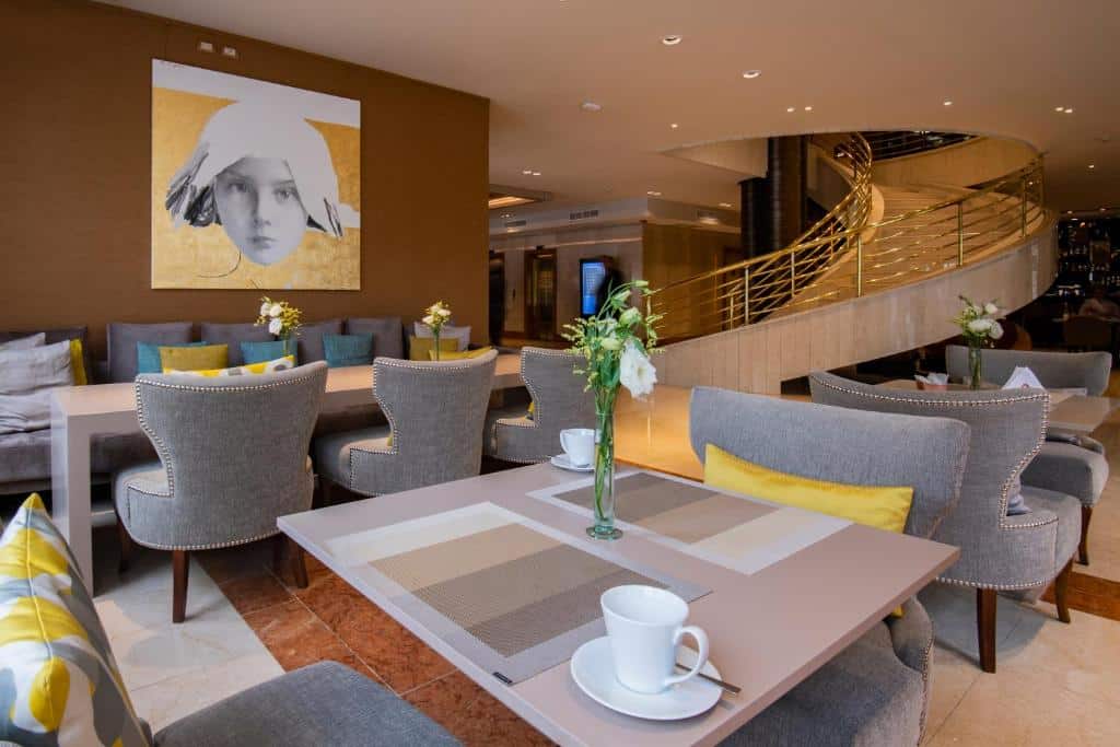 sala de refeições com mesas quadradas e poltronas cinzas que parecem confortáveis no Hotel Grand Brizo Buenos Aires, com uma escada espiralada grande atrás e quadros na parede, tudo combinando em tons de cinza e amarelo