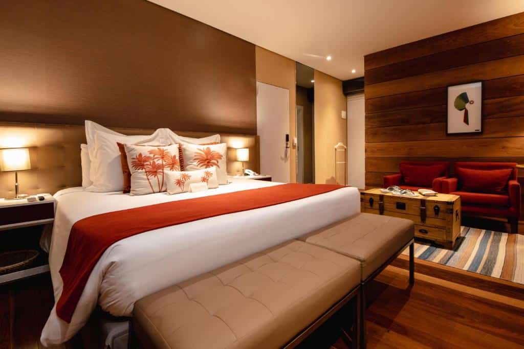 Foto do quarto do Sanma Hotel, ilustrando o post sobre Onde ficar em Foz do Iguaçu. Uma cama box de casal está no centro à esquerda da imagem, e há um banco com almofadas no pé da cama. Na direita do quarto há duas poltronas vermelhas e uma mesa de centro em estilo de baú.