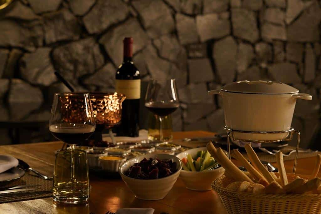 Foto do Sanma Hotel, mostrando uma mesa com aperitivos, fondue, uma garrafa de vinho e duas taças. A parede no fundo é de pedra.