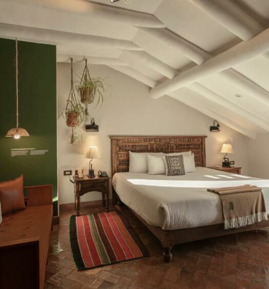 quarto do Antigua Casona San Blas, um dos hotéis em Cusco, com uma cama de casal, uma banqueta com almofadas e um tapete colorido. O teto é inclinado para a direita, dando a sensação de um espaço mais reservado