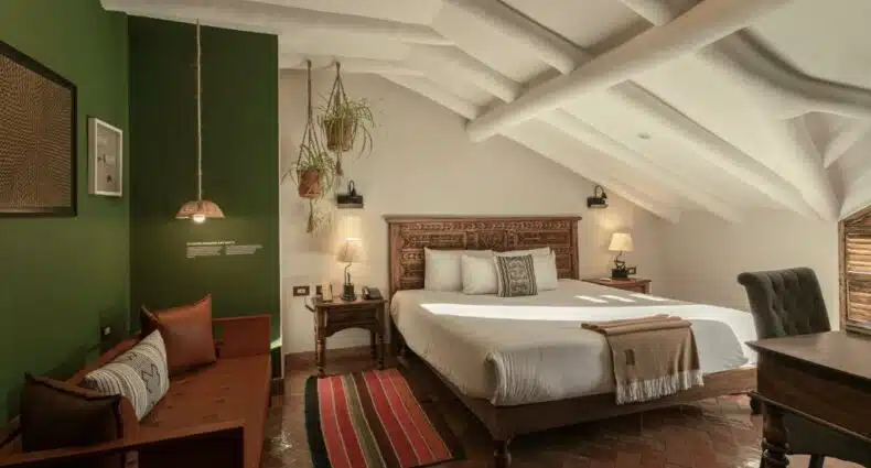 quarto do Antigua Casona San Blas, no Peru, um dos hotéis em Cusco, com uma cama de casal, uma banqueta com almofadas e um tapete colorido. O teto é inclinado para a direita, dando a sensação de um espaço mais reservado