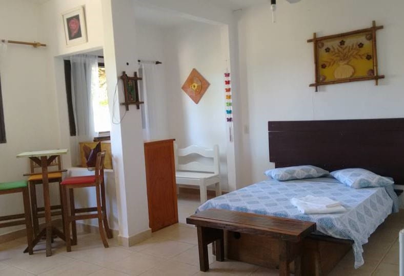 Quarto da Suites Brilho do Sol. Uma cama de casal está no canto direito com um banco no pé. Do lado esquerdo estão uum banco, um armário e algumas cadeiras.