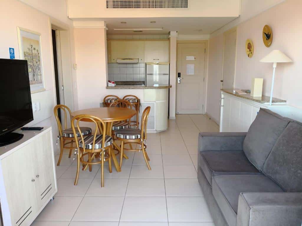 Sala e cozinha do airbnb Suítes e Studios no Hotel Jurerê Beach Village. A direita está um sofá de dois lugares, posicionado em frente a televisão, que fica em cima de um pequeno rack. Ao fundo é possível ver a cozinha e a porta de entrada. A cozinha possui uma bancada com duas cadeiras, em frente a essa bancada está uma mesa redonda com quatro cadeiras.