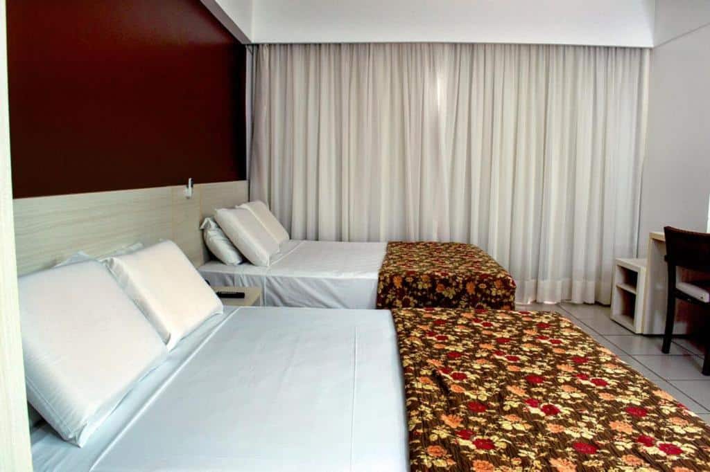 Quarto do Suites Le Jardin. Uma cama de casal e uma cama de solteiro do lado esquerdo, de frente uma mesa de trabalho, no fundo uma parede com cortinas fechadas.