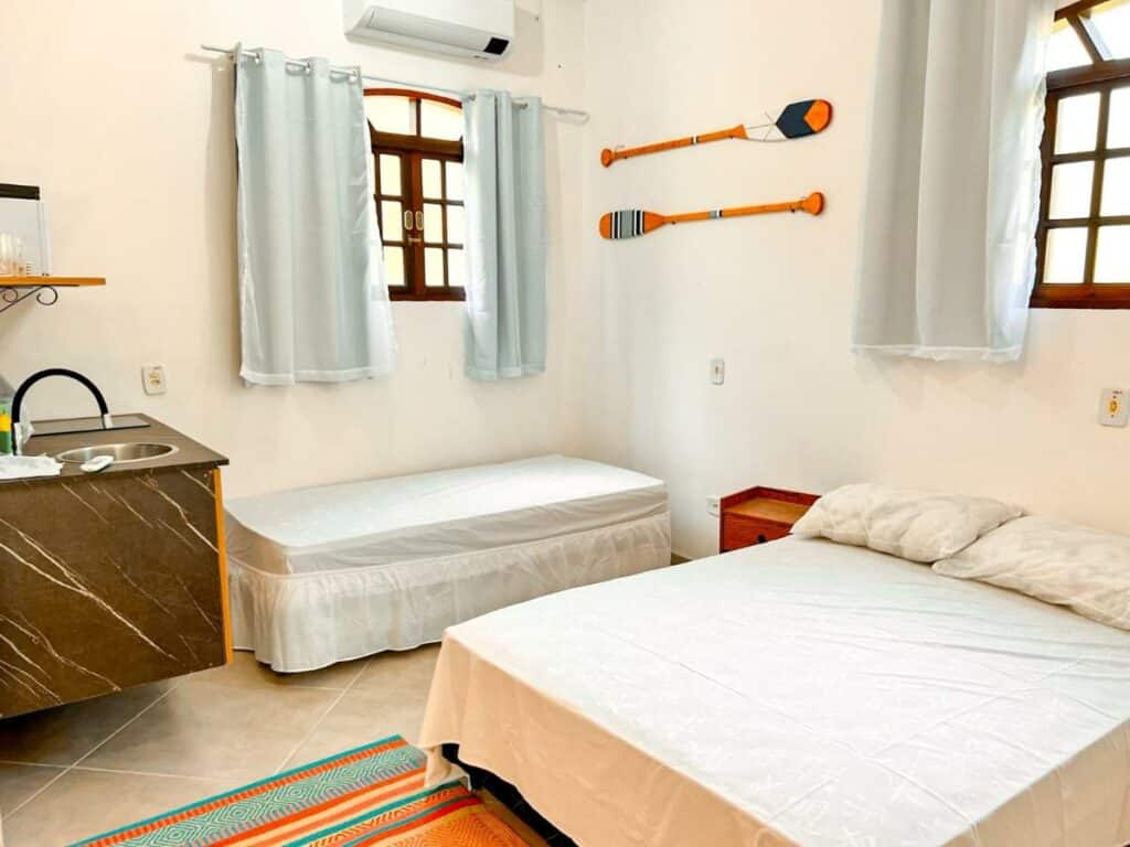 Quarto das Suítes Naturaleza, um dos airbnb em Ilhabela. Uma cama de casal e uma de solteiro tem uma mesinha de cabeceira entre si. Aos pés da cama de solteiro fica a pia da cozinha compacta com cooktop. Há uma janela em cada uma das paredes.