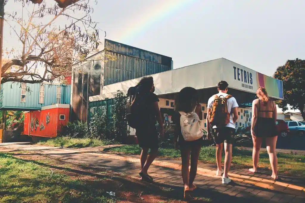 Quatro jovens caminhando em direção ao Tetris Container Hostel. Vemos os vários containers da hospedagem e uma fachada com o nome dela.