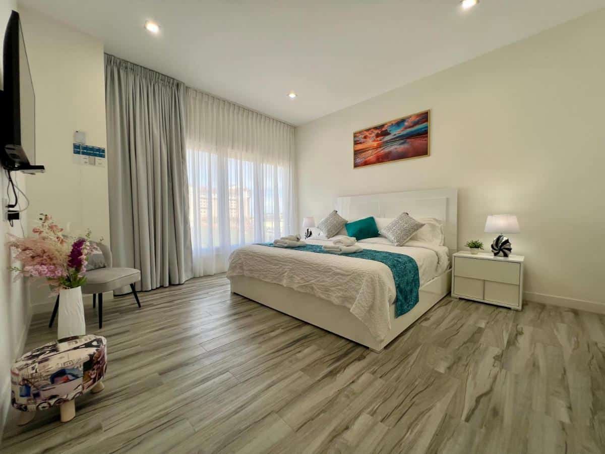 Quarto de um dos apartamentos do The Cove - Condo Hotel. Uma cama de casal está no meio do ambiente, encostada na parede. Ao seu lado direito está uma mesinha com abajur e ao lado esquerdo uma grande janela com cortina. Em frente a cama está uma tv na parede, uma cadeira e um vaso de flor.