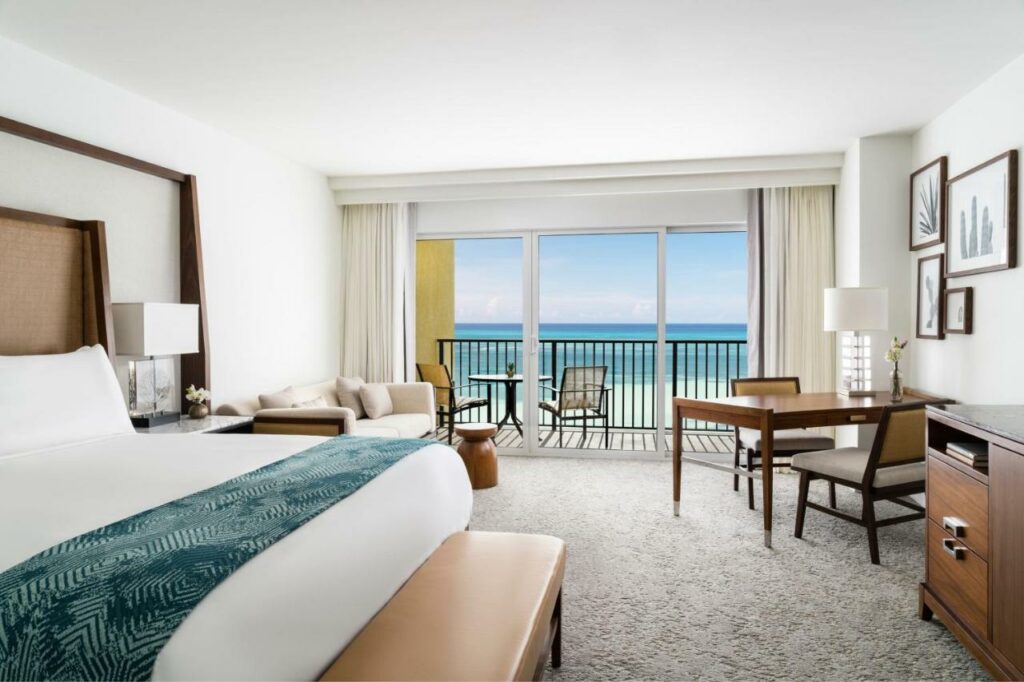 Quarto do The Ritz-Carlton com cama de casal do lado esquerdo da imagem no pé da cama um banco e do lado esquerdo da cama um sofá e em frente ao sofá uma mesa de madeira com duas cadeiras. Representa Aruba.
