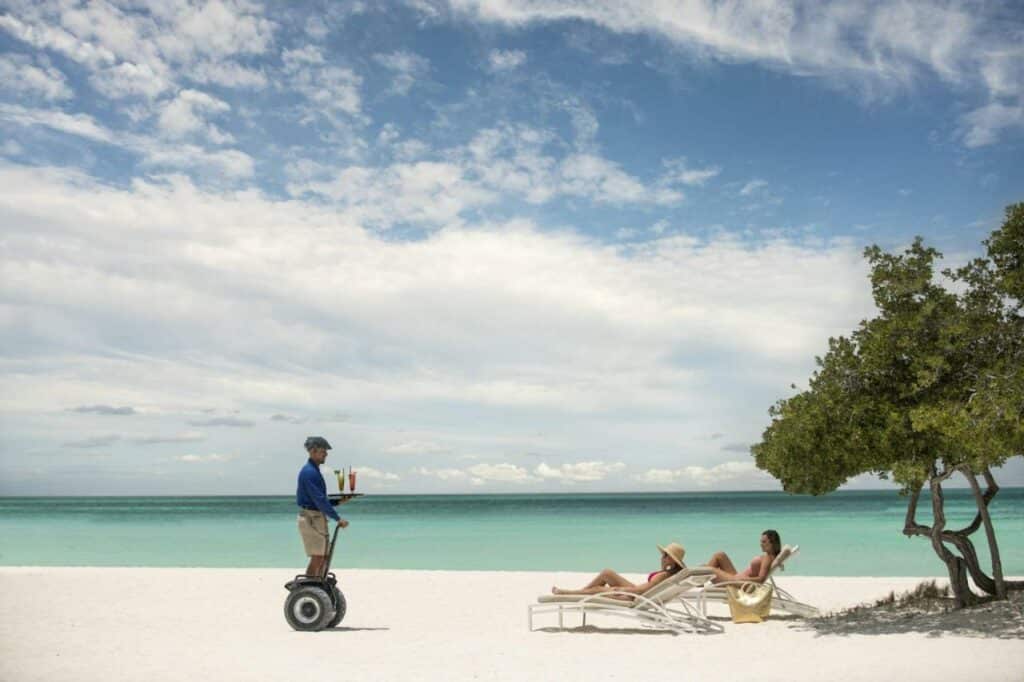 Na imagem um funcionário do The Ritz-Carlton, Aruba está levando bebidas para duas pessoas deitadas em espreguiçadeiras na areia da praia, perto do mar. O The Ritz é um dos hotéis de luxo em Aruba.