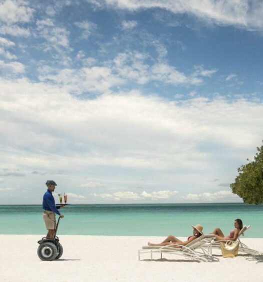 Na imagem um funcionário do The Ritz-Carlton, Aruba está levando bebidas para duas pessoas deitadas em espreguiçadeiras na areia da praia, perto do mar. O The Ritz é um dos hotéis de luxo em Aruba.