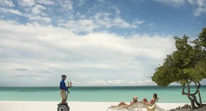 Na imagem um funcionário do The Ritz-Carlton, Aruba está levando bebidas para duas pessoas deitadas em espreguiçadeiras na areia da praia, perto do mar. O The Ritz é um dos hotéis de luxo em Aruba.