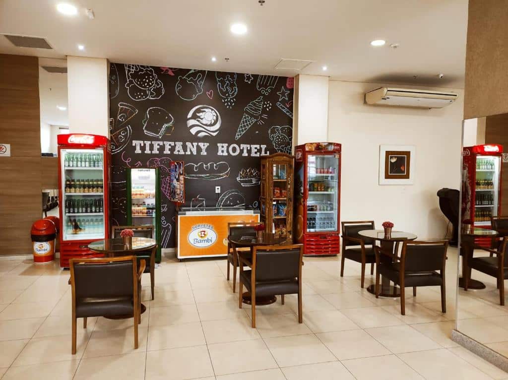 Área de refeições do Tiffany Hotel com mesas e cadeiras estofadas, refrigeradores com bebidas, uma prateleira com salgadinhas e um refrigerador com sorvetes