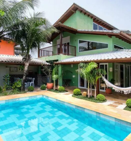 Piscina e vista de fora da casa UBATUBA PRAIA DO SAPE. A piscina é grande, retangular e está no meio. Ao redor estão várias árvores pequenas e uma rede. Ao fundo está a própria casa. Este é um airbnb na Praia do Sapê.