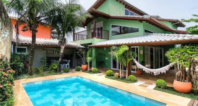 Piscina e vista de fora da casa UBATUBA PRAIA DO SAPE. A piscina é grande, retangular e está no meio. Ao redor estão várias árvores pequenas e uma rede. Ao fundo está a própria casa. Este é um airbnb na Praia do Sapê.
