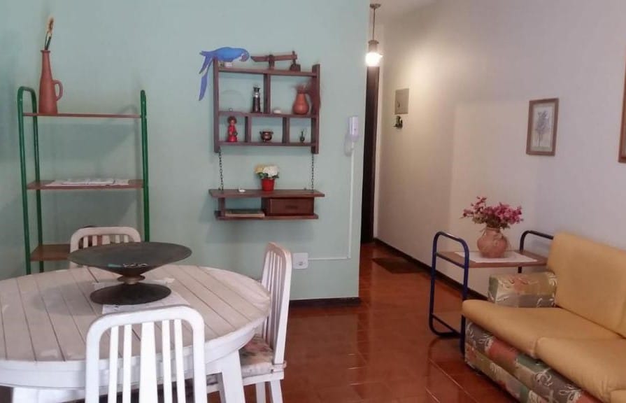 Sala de estar do Ubatuba Tenório apto térreo 6 pess. No canto direito há um sofá encostado na parede e ao lado de uma mesinha com um jarro de flor. Em frente a ele está uma mesa redonda de quatro lugares. Na parede do outro lado estão algumas prateleiras.