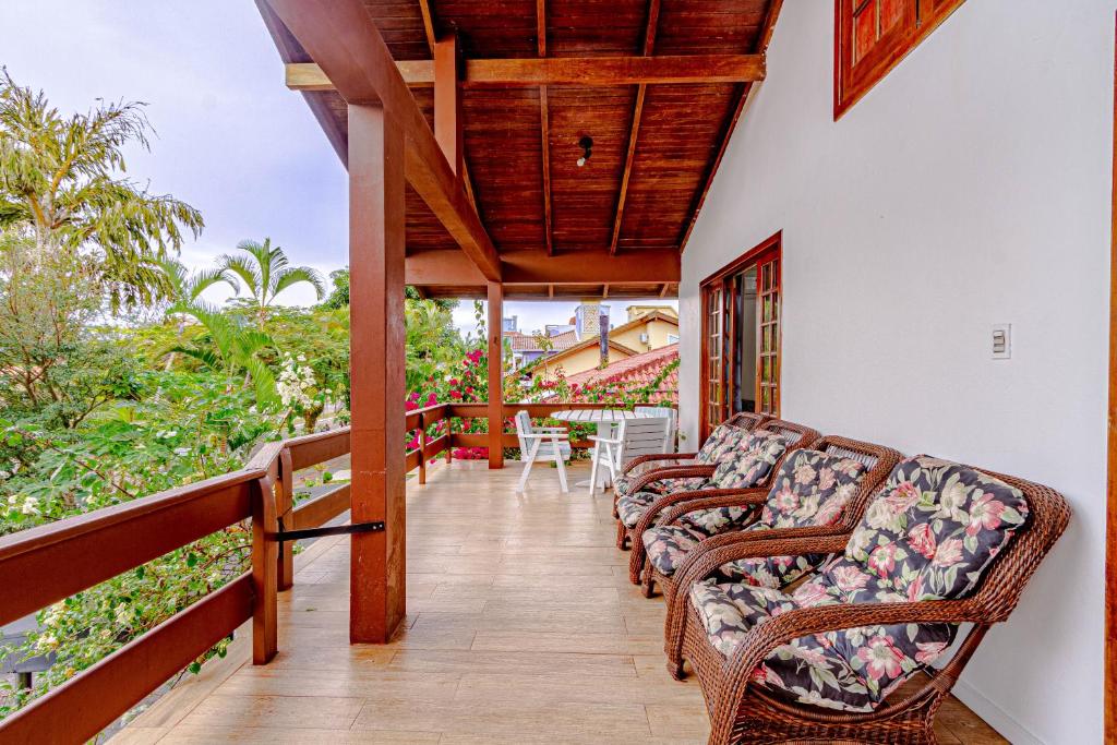Varanda do airbnb Suítes Jurerê Internacional. No lado direito estão quatro cadeiras acolchoadas, no fundo há uma porta de madeira que leva a área interna do quarto e uma mesa redonda com três cadeiras. Em frente ao parapeito, que cerca toda a varanda, há muitas árvores, flores e plantas.